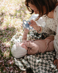 Woman and baby with mushie Daisy Rattle Teether among pink petals on a blanket outdoors.