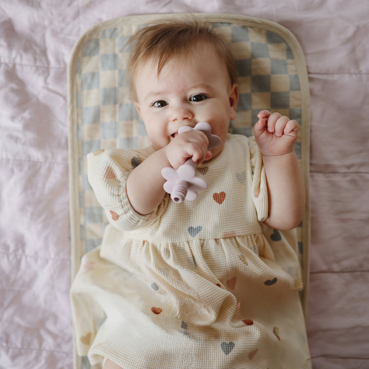 A baby chews a flower-shaped teether while lying on a Mushie Changing Pad Liner.