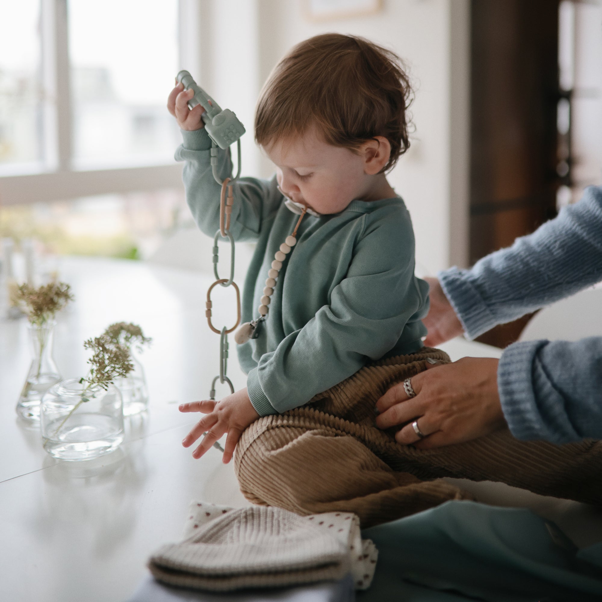 A toddler uses a FRIGG Butterfly Anatomical Silicone Pacifier (0-6 Months) while sitting at a table, holding a toy. An adult supports the child in a cozy, softly lit room decorated with vases of flowers and folded clothes.