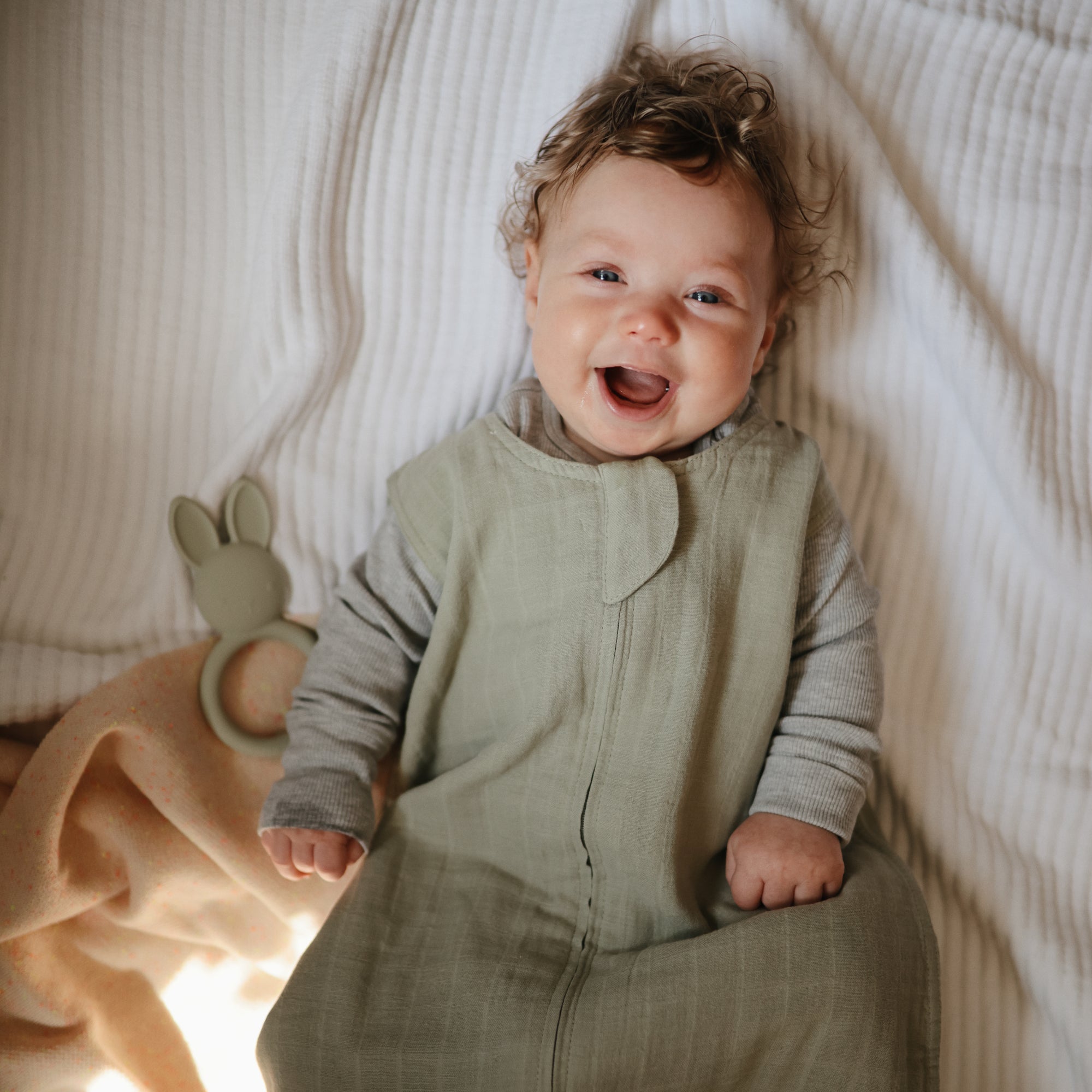Smiling baby in green outfit on bed with mushie Bunny Teether and cozy blanket nearby.
