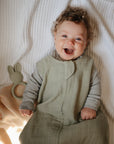 Smiling baby in green outfit on bed with mushie Bunny Teether and cozy blanket nearby.