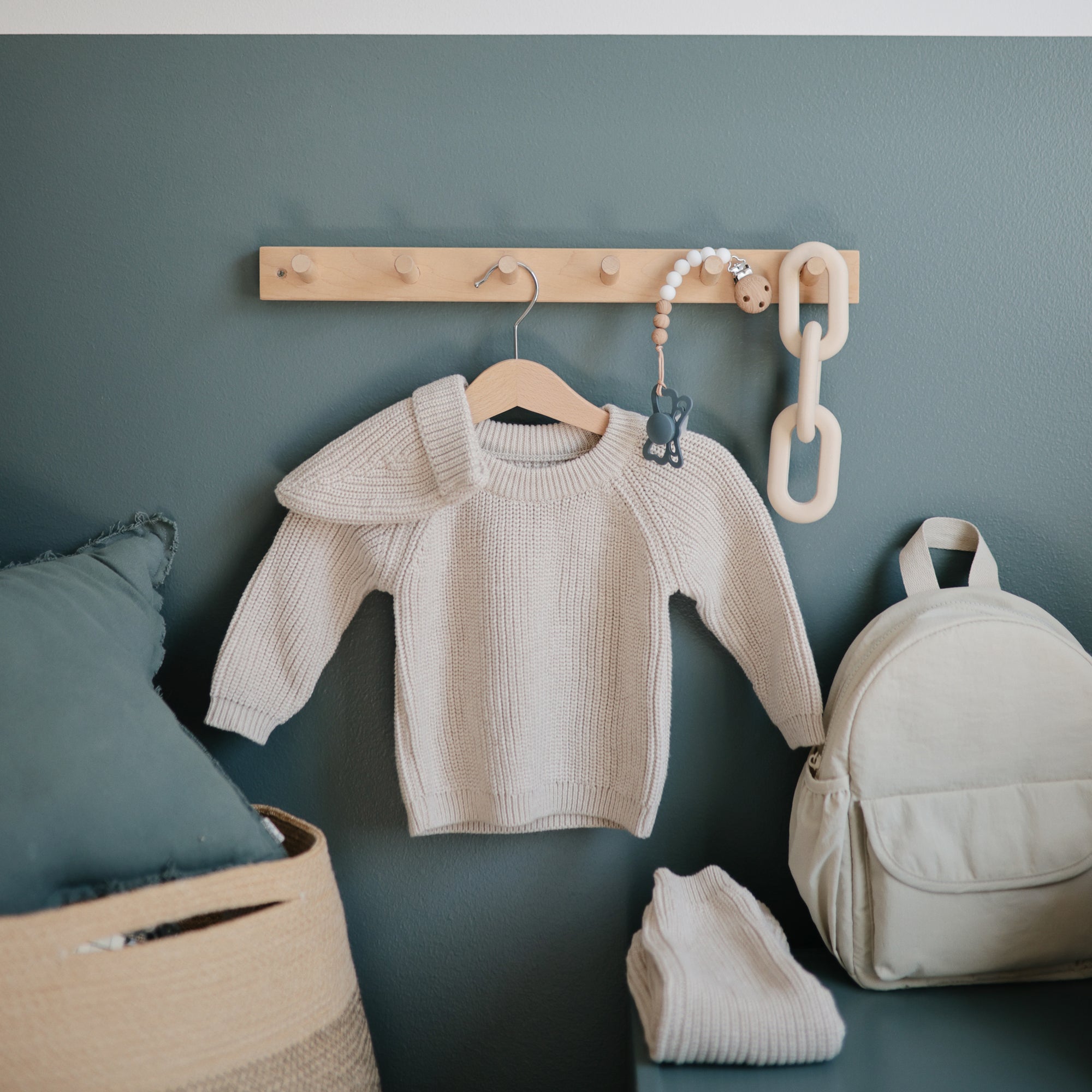 A small beige knit sweater hangs on a wooden peg rack against a sage green wall, with a folded matching garment, beige backpack, basket, and FRIGG Butterfly Anatomical Silicone Pacifier 2-Pack (0-6 Months) nearby. Large chain links hang from the rack.
