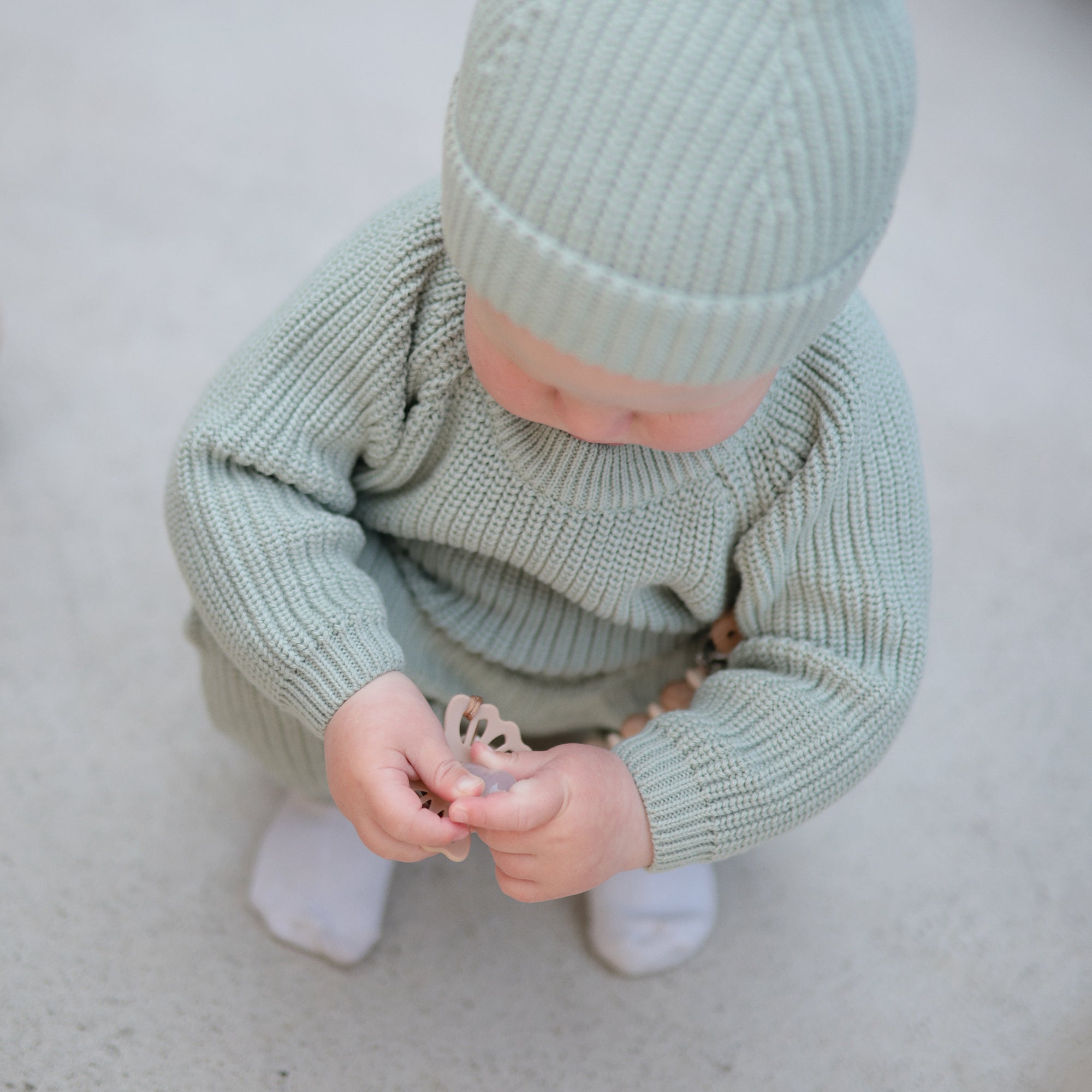 A baby in a light green knit outfit and hat squats on a light surface, playing with a FRIGG Lucky Symmetrical Silicone Pacifier (0-6 Months) in both hands. The white socks on the baby's feet are visible.