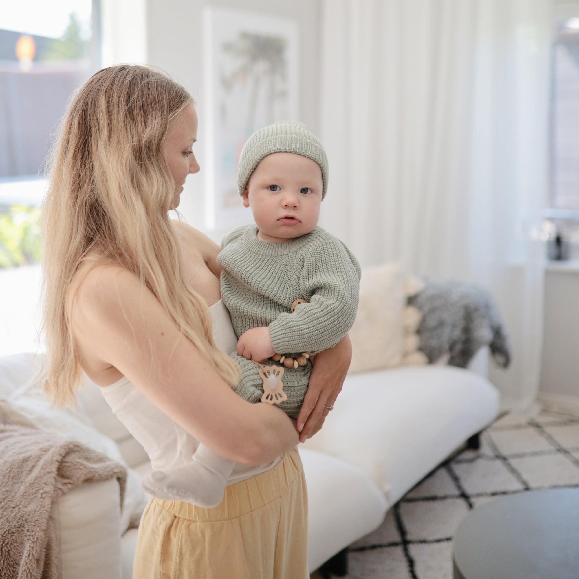 A woman with long blonde hair holds a baby in a light green knit outfit and hat, soothing the infant with FRIGG Butterfly Anatomical Silicone Pacifiers (0-6 Months), in a bright living room filled with white furniture and soft natural light.