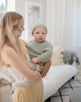 A woman with long blonde hair holds a baby in a light green knit outfit and hat, soothing the infant with FRIGG Butterfly Anatomical Silicone Pacifiers (0-6 Months), in a bright living room filled with white furniture and soft natural light.