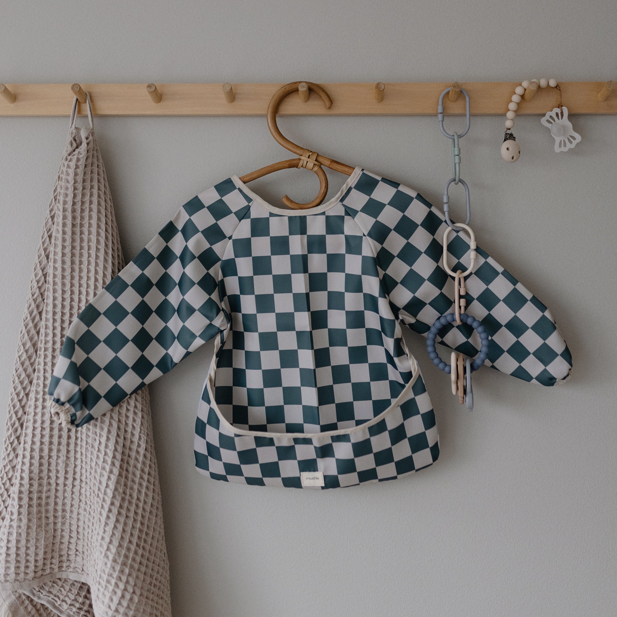 A blue and white checkered long-sleeve bib hangs on a wooden peg rack beside a beige textured towel and a chain featuring FRIGG Lucky Symmetrical Silicone Pacifiers (0-6 Months) for baby comfort and safety.