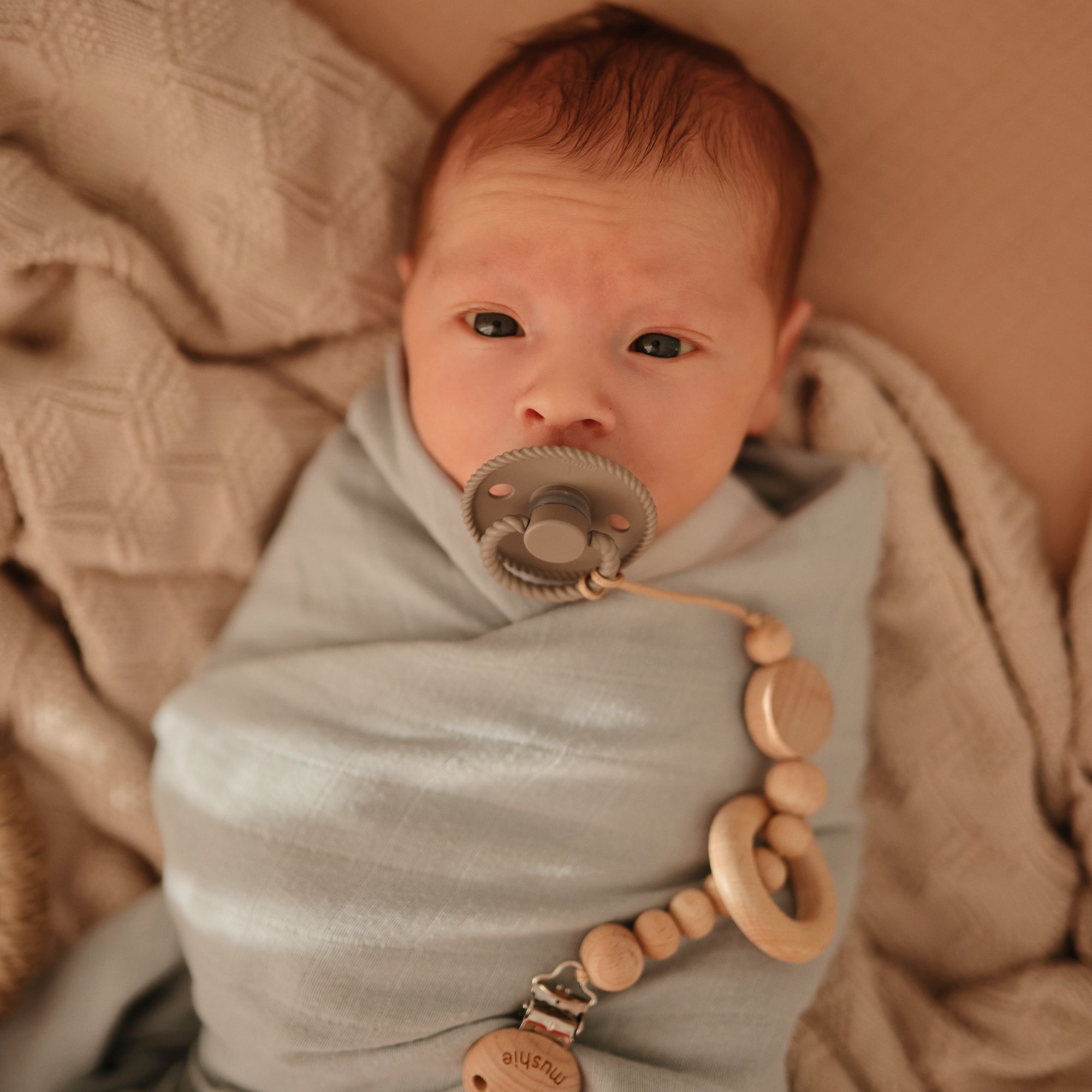 A newborn with light skin and reddish hair is swaddled in a light blue blanket on a beige surface, using a FRIGG Rope Silicone Pacifier (2-Pack) clipped to the blanket with a wooden beaded chain.