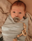 A newborn with light skin and reddish hair is swaddled in a light blue blanket on a beige surface, using a FRIGG Rope Silicone Pacifier (2-Pack) clipped to the blanket with a wooden beaded chain.