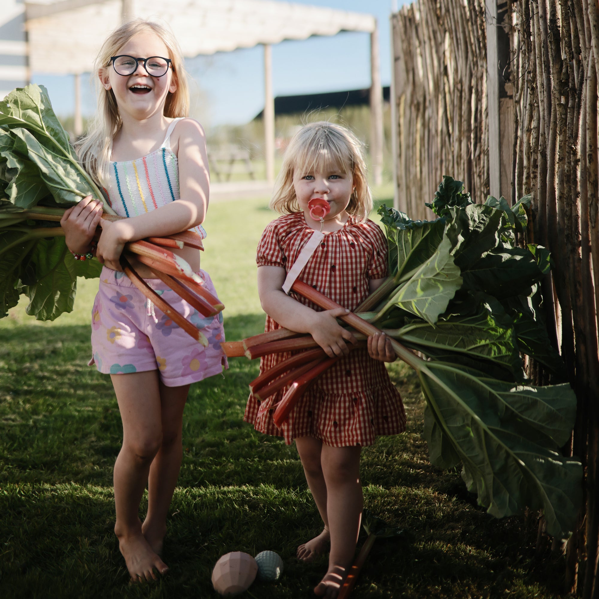 Two girls outdoors holding rhubarb leaves and mushie Linen Pacifier Clip 2-Pack on the grass.