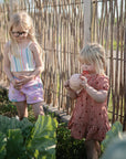 Two girls in a garden; one enjoys fruit holding a mushie Nesting Spheres Sensory Toy.