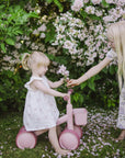 Two blonde girls share flowers by a mushie Mini Toddler Bike in a lush garden.
