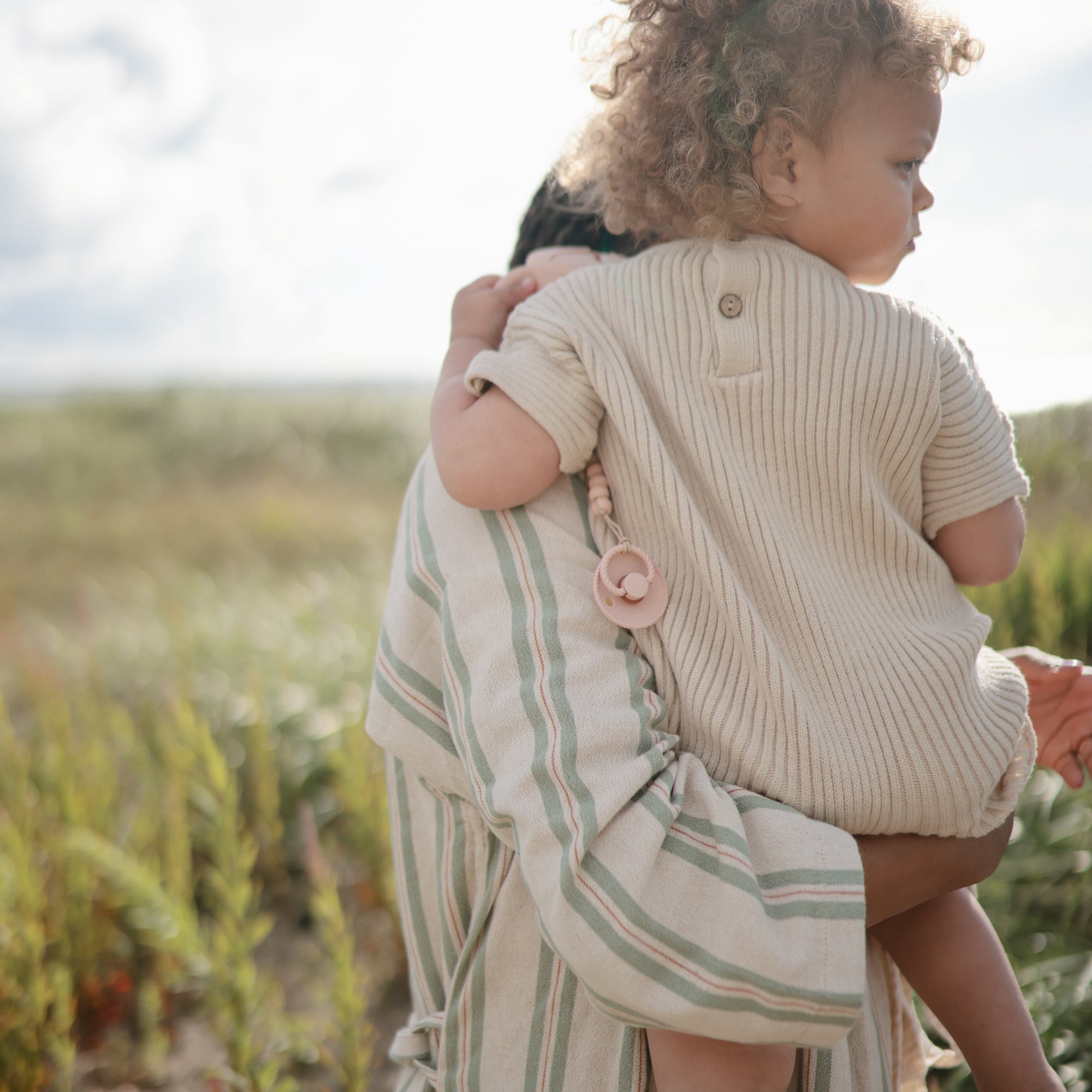 Outdoors, an adult in stripes carries a curly-haired toddler in a beige romper. The toddler holds a Mushie FRIGG Rope Natural Rubber Pacifier, clipped to their outfit, with greenery and sunlight in the background.