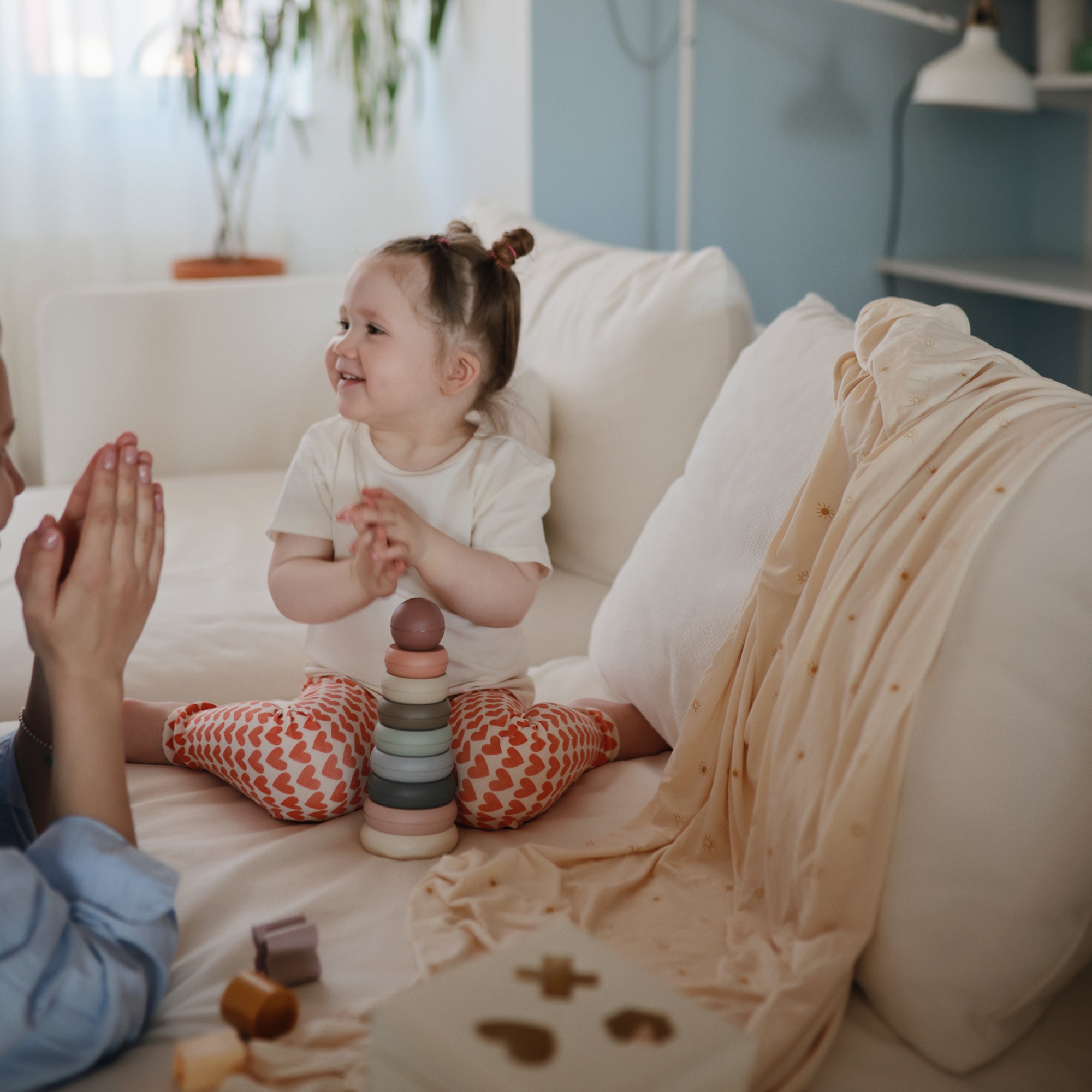 A young child sits on a white couch, smiling beside an adult. Nearby are colorful stacking toys and a shape sorter. A mushie Stretchy Swaddle is draped in the softly lit room, with a plant and other blankets in the background.