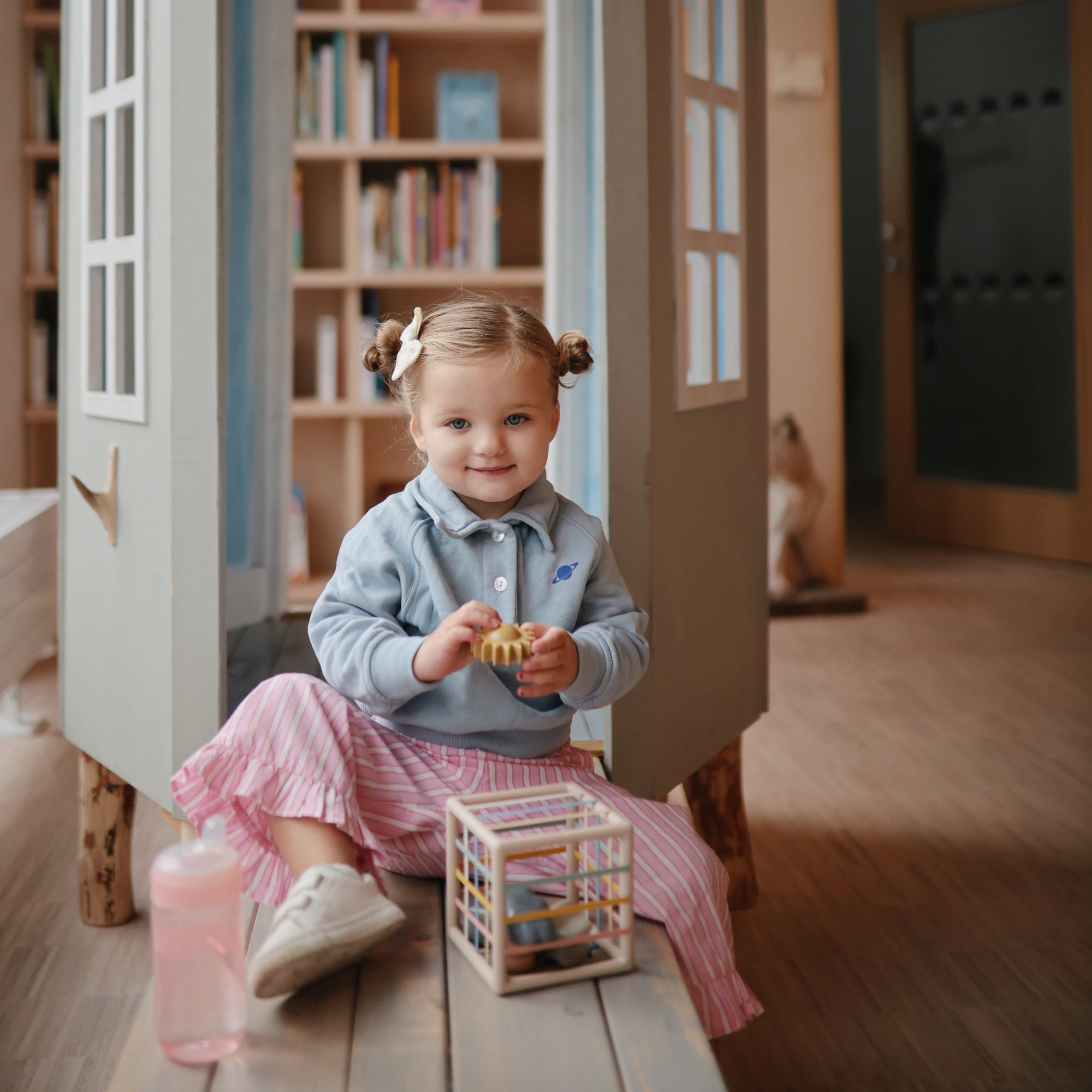 Smiling girl on bench holds mushie Elastic Shape Sorter, with bookshelves in the background.