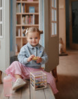 Smiling girl on bench holds mushie Elastic Shape Sorter, with bookshelves in the background.