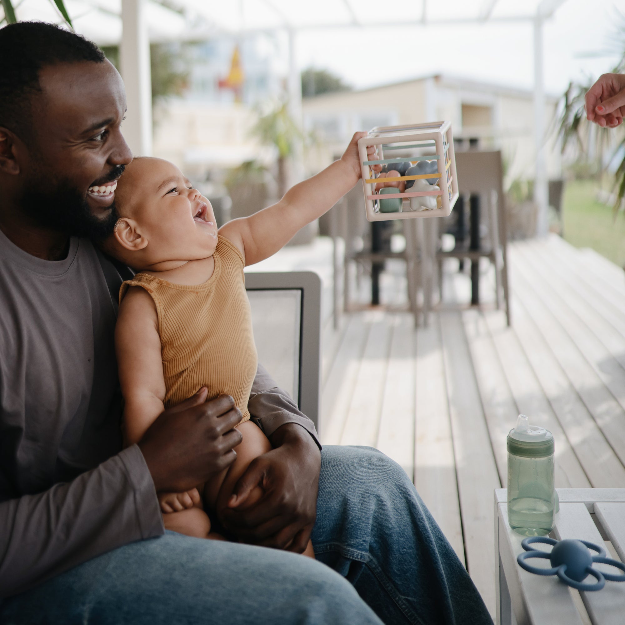 Smiling man with baby reaches for mushie Elastic Shape Sorter at an outdoor patio table.