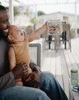 Smiling man with baby reaches for mushie Elastic Shape Sorter at an outdoor patio table.