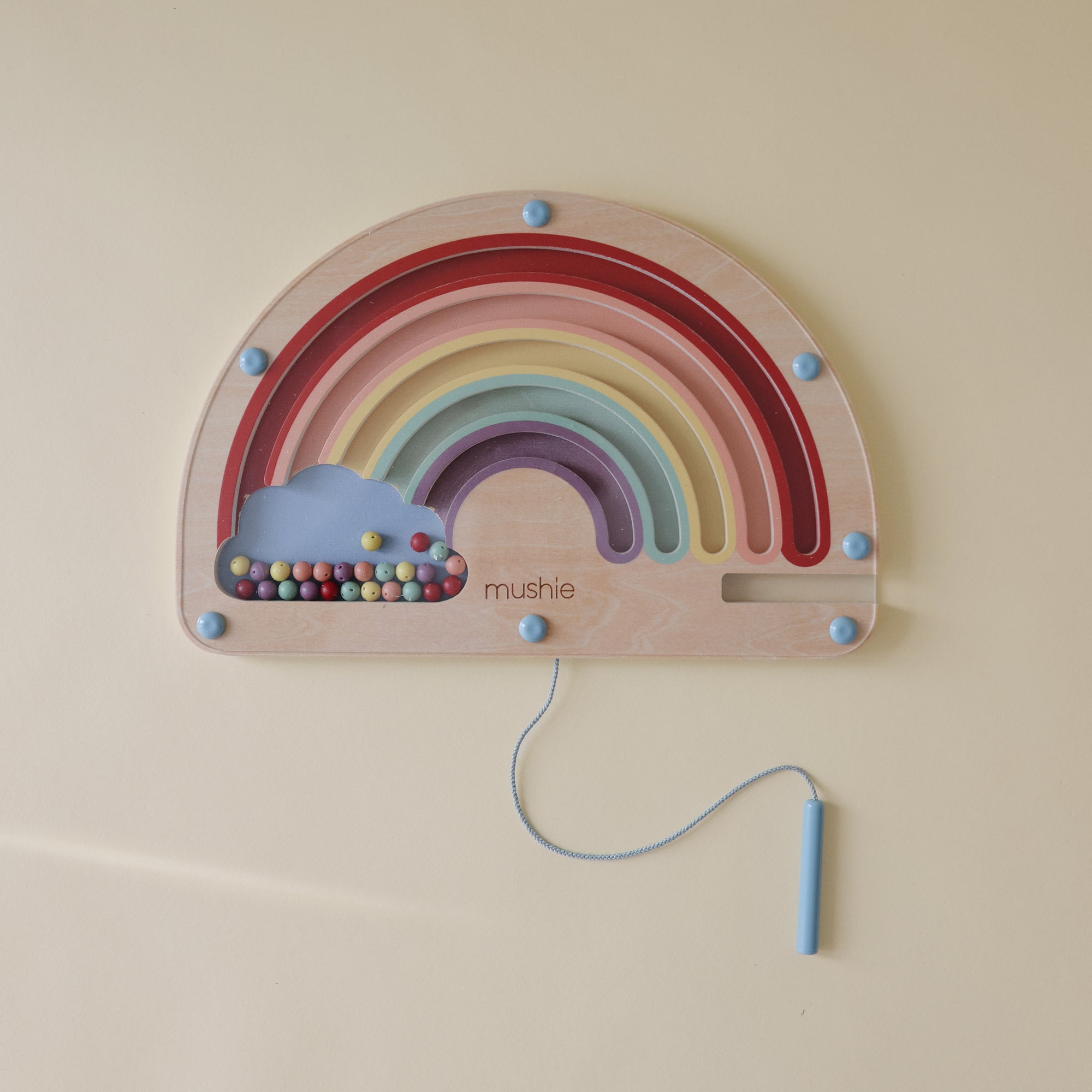 The mushie Wooden Magnetic Rainbow Maze features colorful arches, a blue cloud, and pastel beads. Kids use a handled blue string to guide beads on the pale yellow surface, encouraging fine motor skills and screen-free play.