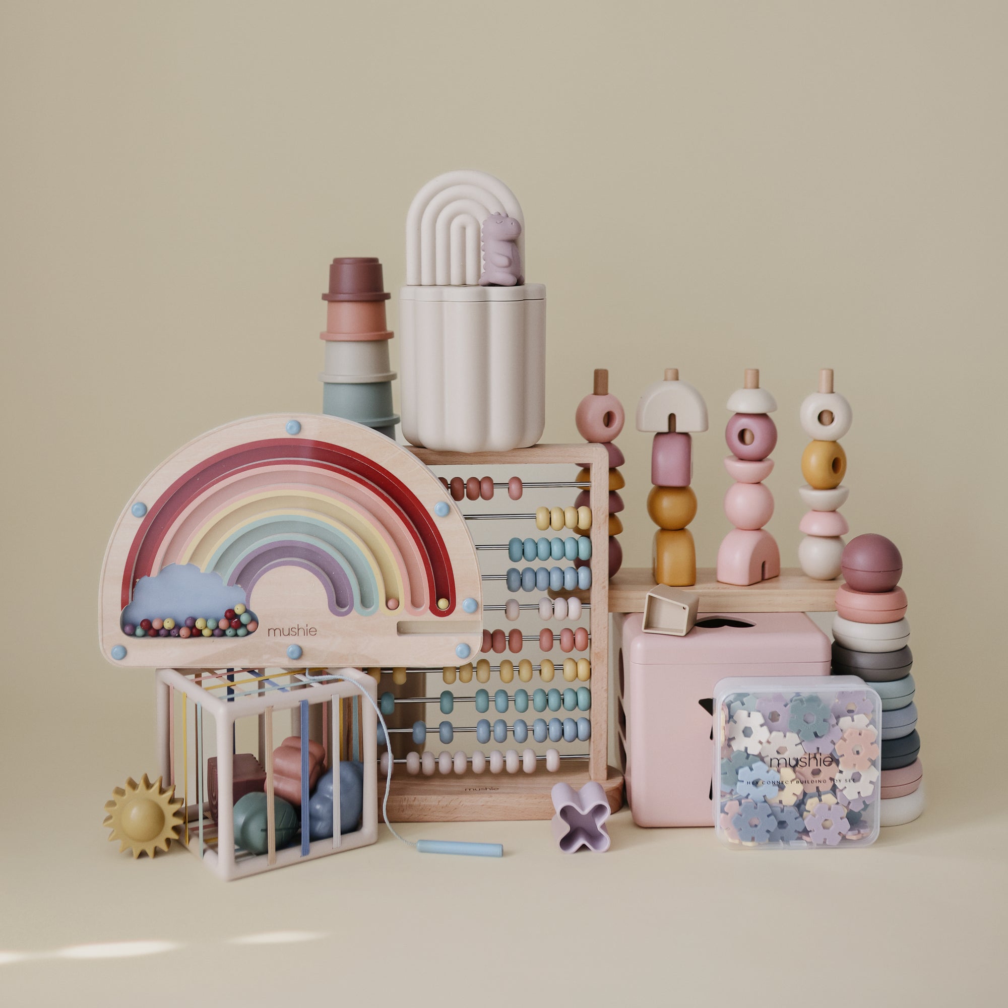 A pastel display of children’s toys, featuring the mushie Wooden Magnetic Rainbow Maze, stacking rings, abacuses, beads, and geometric shapes—all perfect for screen-free fun and developing fine motor skills.
