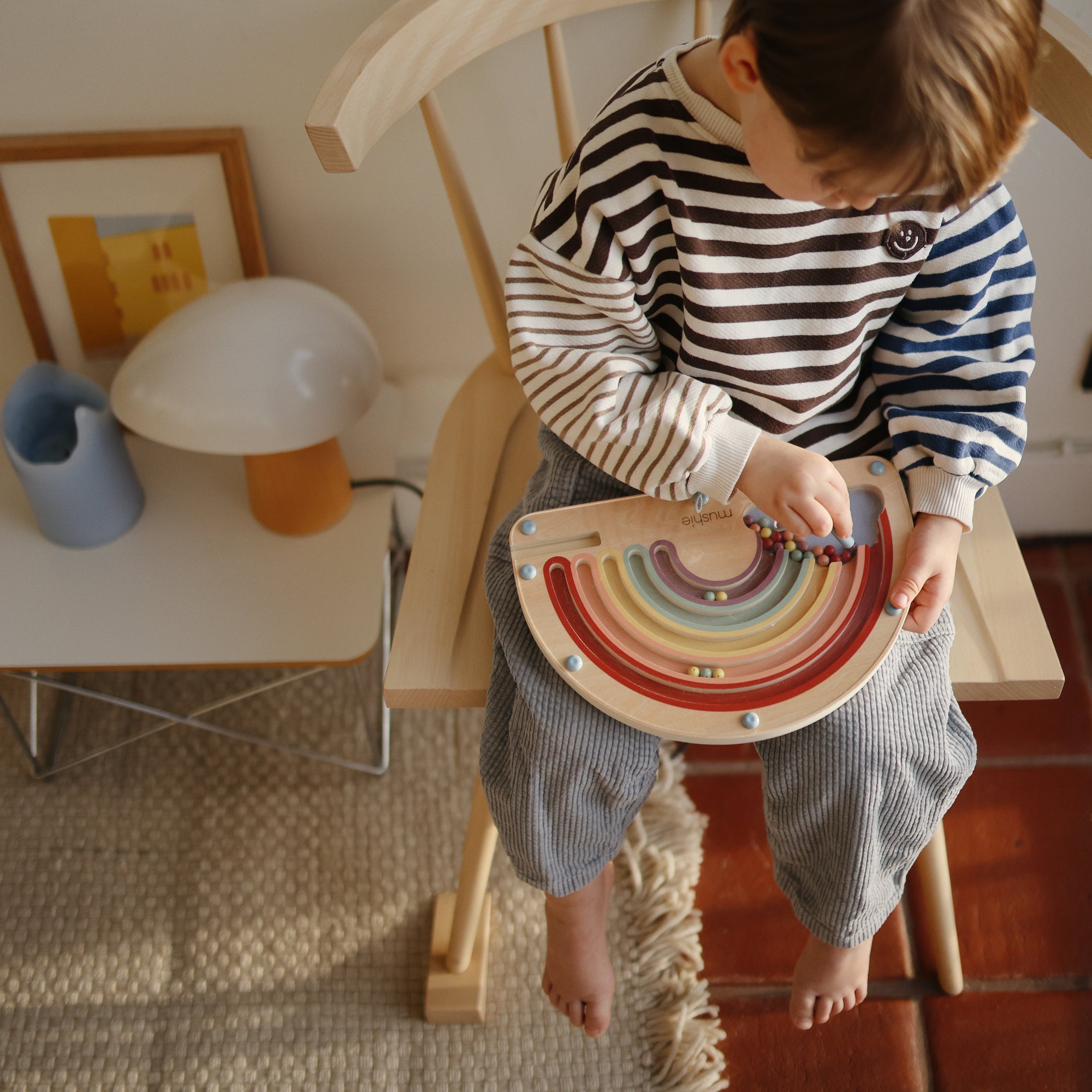 A barefoot child in a striped shirt plays with the mushie Wooden Magnetic Rainbow Maze, enjoying screen-free fun that builds fine motor skills. On a nearby table are a lamp, blue pitcher, and framed art.