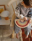 A barefoot child in a striped shirt plays with the mushie Wooden Magnetic Rainbow Maze, enjoying screen-free fun that builds fine motor skills. On a nearby table are a lamp, blue pitcher, and framed art.