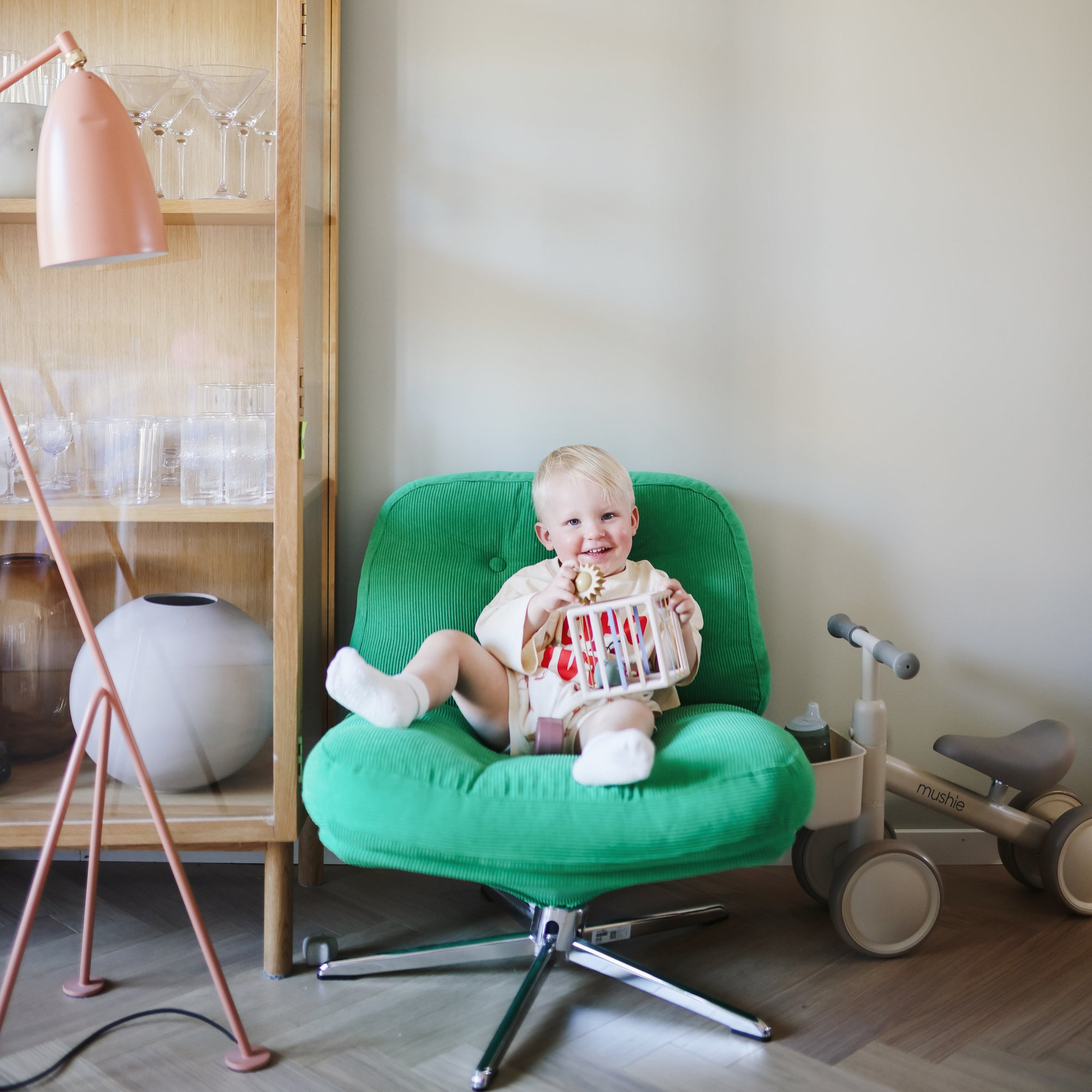 Smiling baby plays with mushie Elastic Shape Sorter on green chair near tricycle and cabinet.