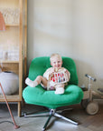 Smiling baby plays with mushie Elastic Shape Sorter on green chair near tricycle and cabinet.
