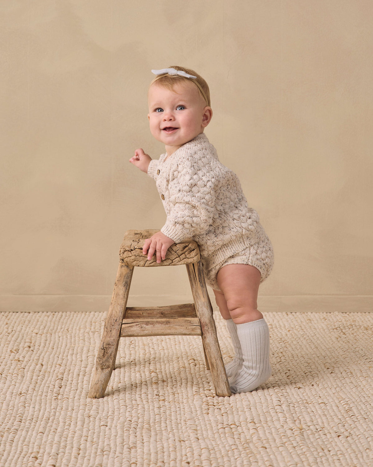 A baby in Rylee + Cru Knit Bloomer Natural Confetti stands on a rug with a beige background.
