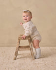 A baby in Rylee + Cru Knit Bloomer Natural Confetti stands on a rug with a beige background.