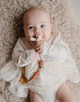 Baby smiles on a soft rug, holding the mushie Figurine Teether.