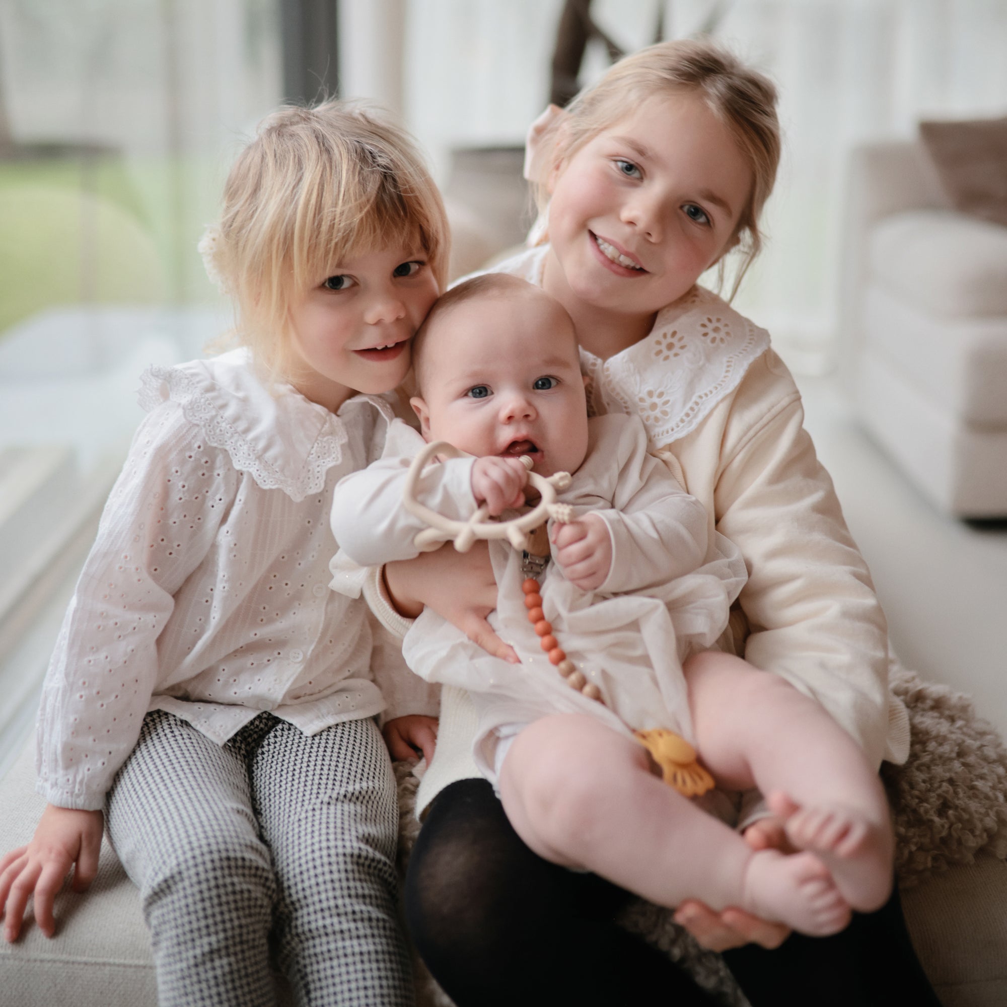 Two older girls on a couch hold a baby in white, who smiles while chewing a FRIGG Lucky Symmetrical Silicone Pacifier (0-6 Months). The heartwarming, cozy scene highlights the comfort and safety of this trusted FRIGG product.