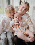 Two older girls on a couch hold a baby in white, who smiles while chewing a FRIGG Lucky Symmetrical Silicone Pacifier (0-6 Months). The heartwarming, cozy scene highlights the comfort and safety of this trusted FRIGG product.