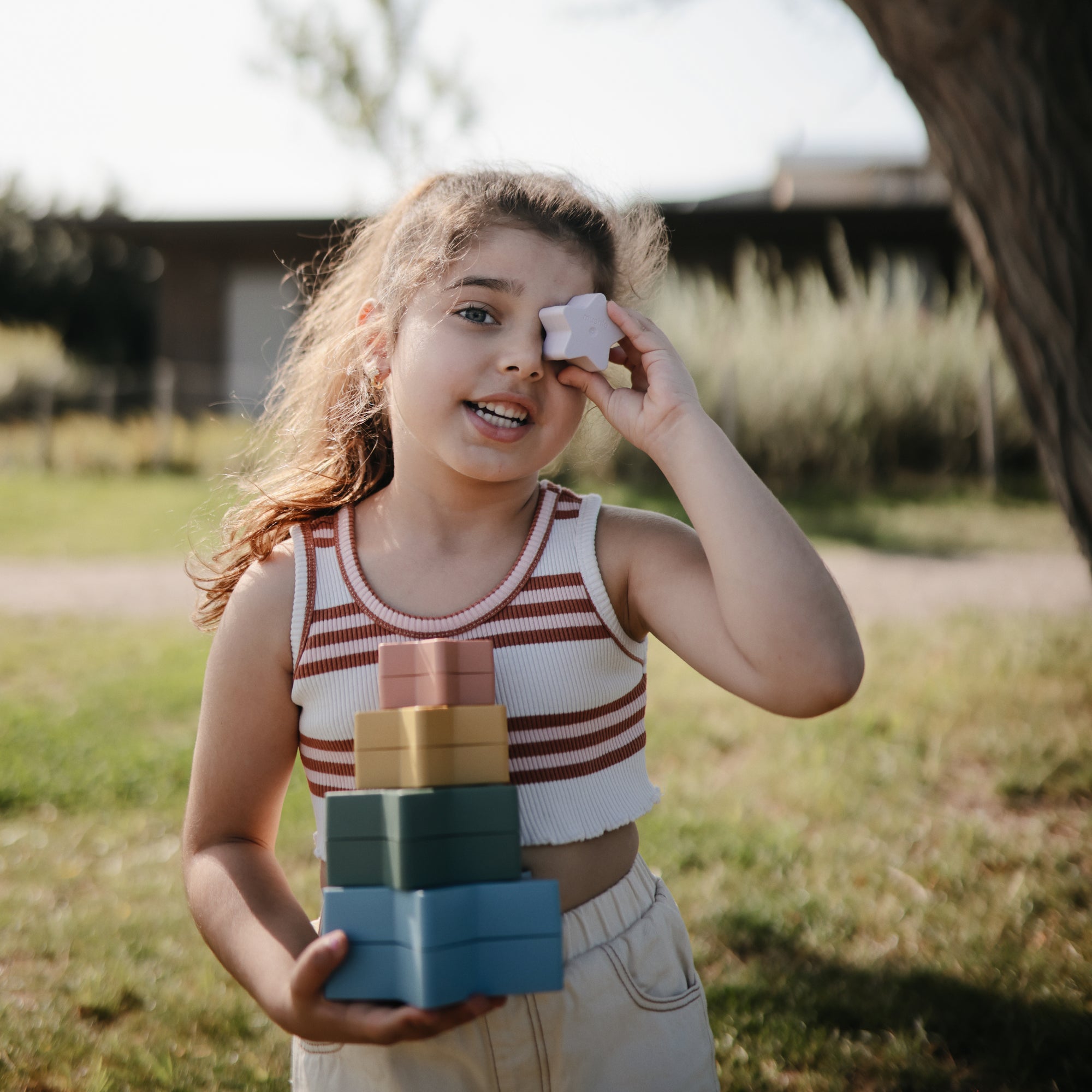 Smiling girl outdoors holds mushie Nesting Stars Toy, stacking a star to her eye.