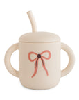 Mushie Silicone Training Cup + Straw—beige, two handles, pink bow; perfect for toddlers.