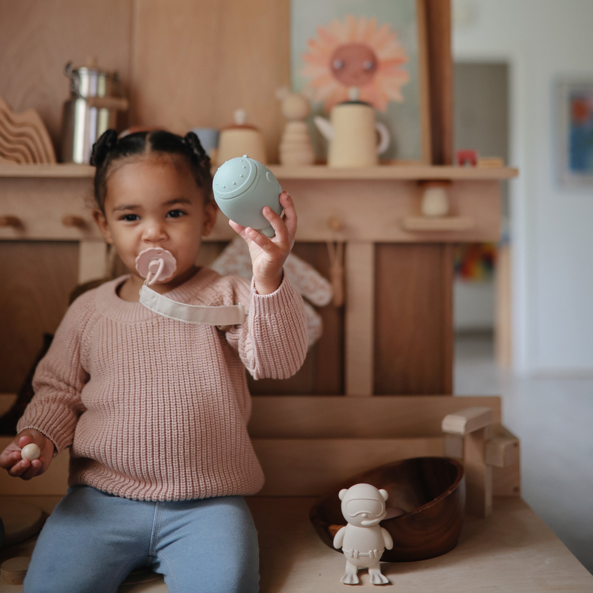 Toddler with pigtails holds a toy and uses a FRIGG Daisy Natural Rubber Pacifier.