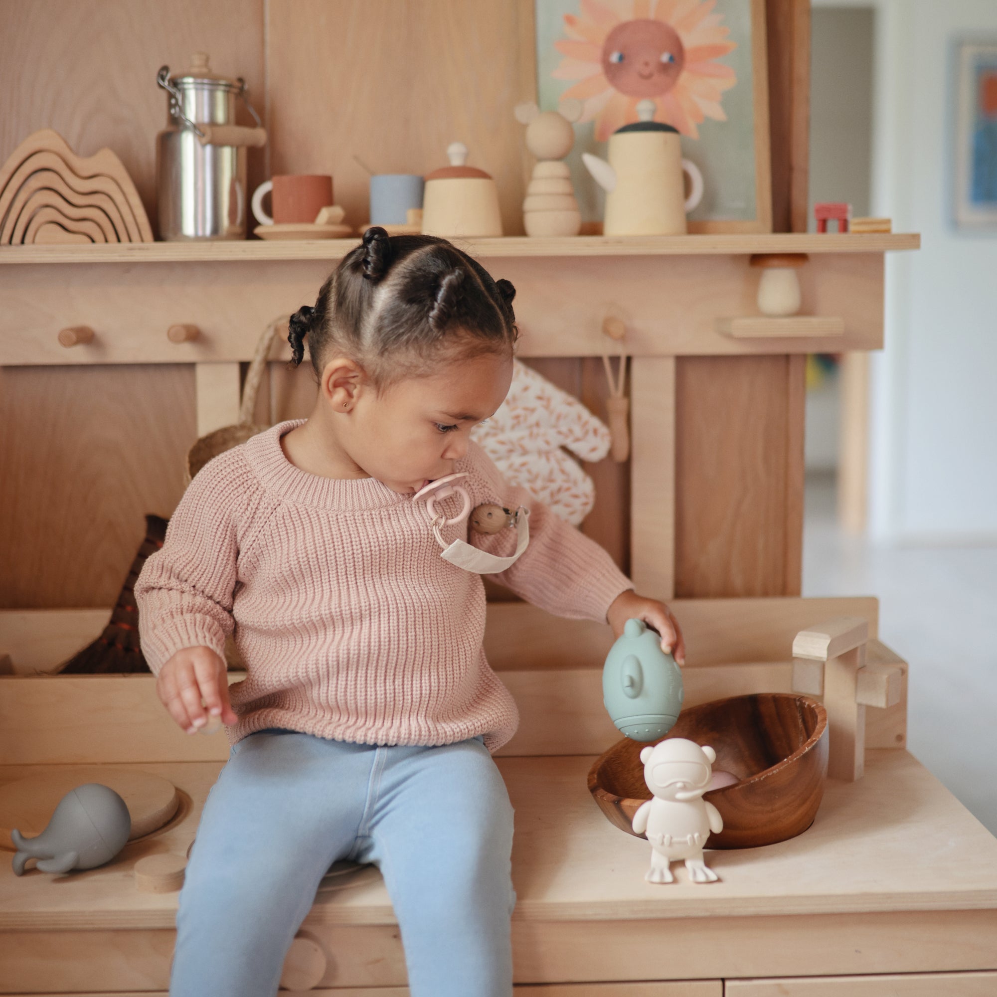 Toddler with wooden toys and a FRIGG Daisy Natural Rubber Pacifier by FRIGG at a play kitchen.