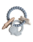 A silicone baby teething toy with beaded blue ring and attached shapes, including a beige rocket, a white star, and a gray astronaut, all linked together on a white background.
