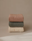 A stack of three blankets against a beige background: mushie Stretchy Swaddle in light brown on top, olive green blanket in the middle, and a cream baby blanket with small sun patterns on the bottom.