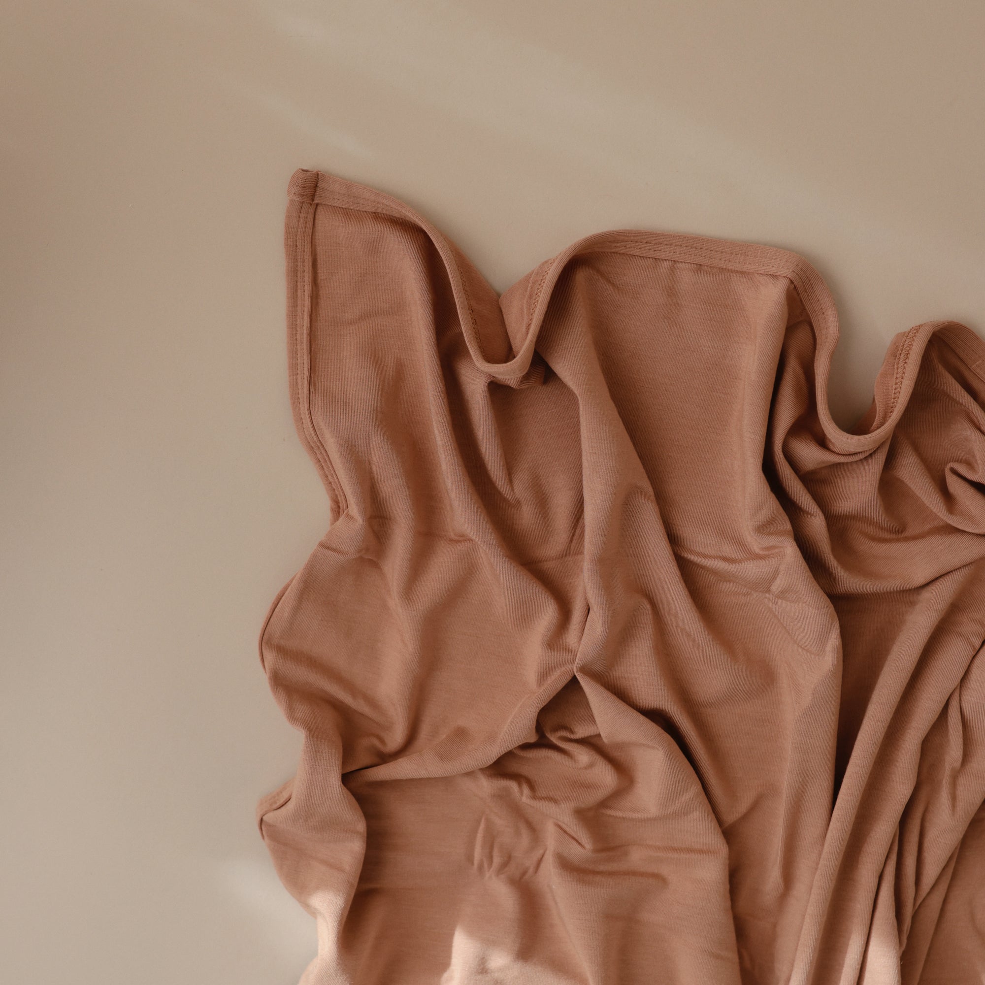 The mushie Stretchy Swaddle in a soft peach shade is draped with gentle folds and shadows on a smooth, light beige surface.