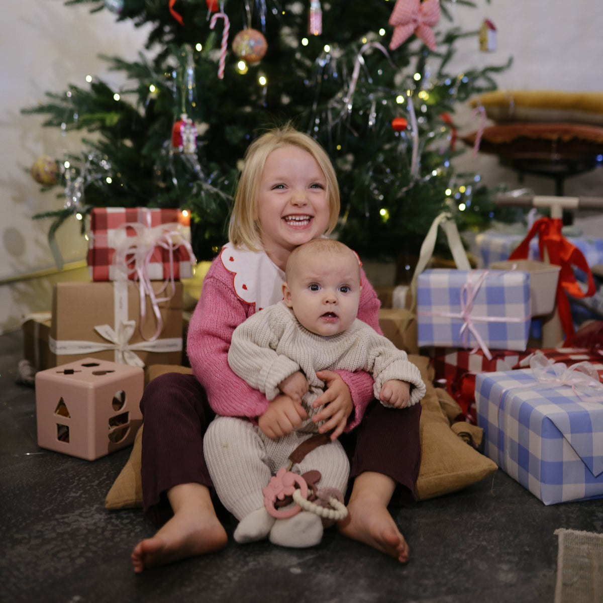 Two kids by a Christmas tree with mushie’s Shape Sorting Box, gifts, and holiday cheer.