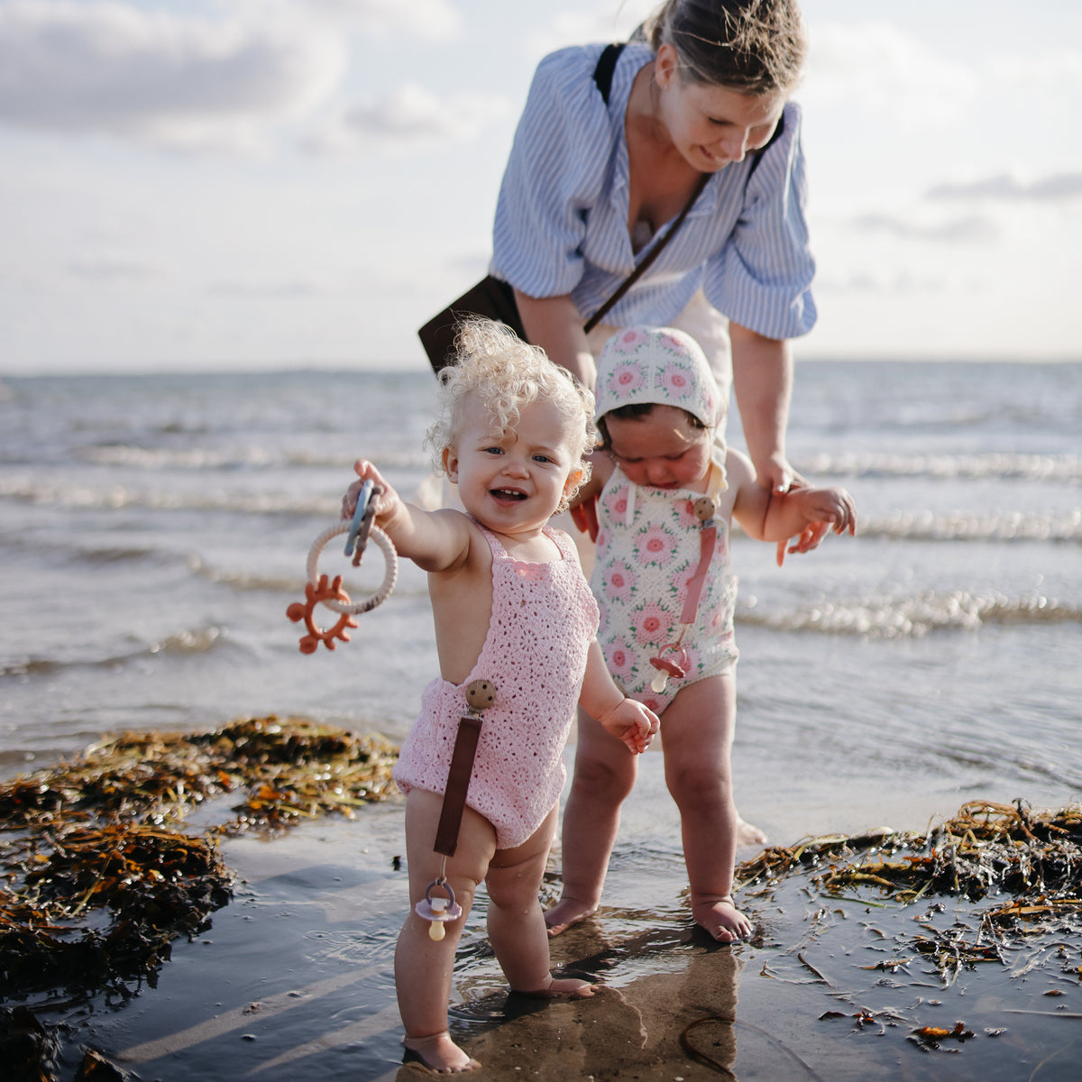 A woman and two toddlers play on the beach, holding mushie Linen Pacifier Clip 2-Pack.