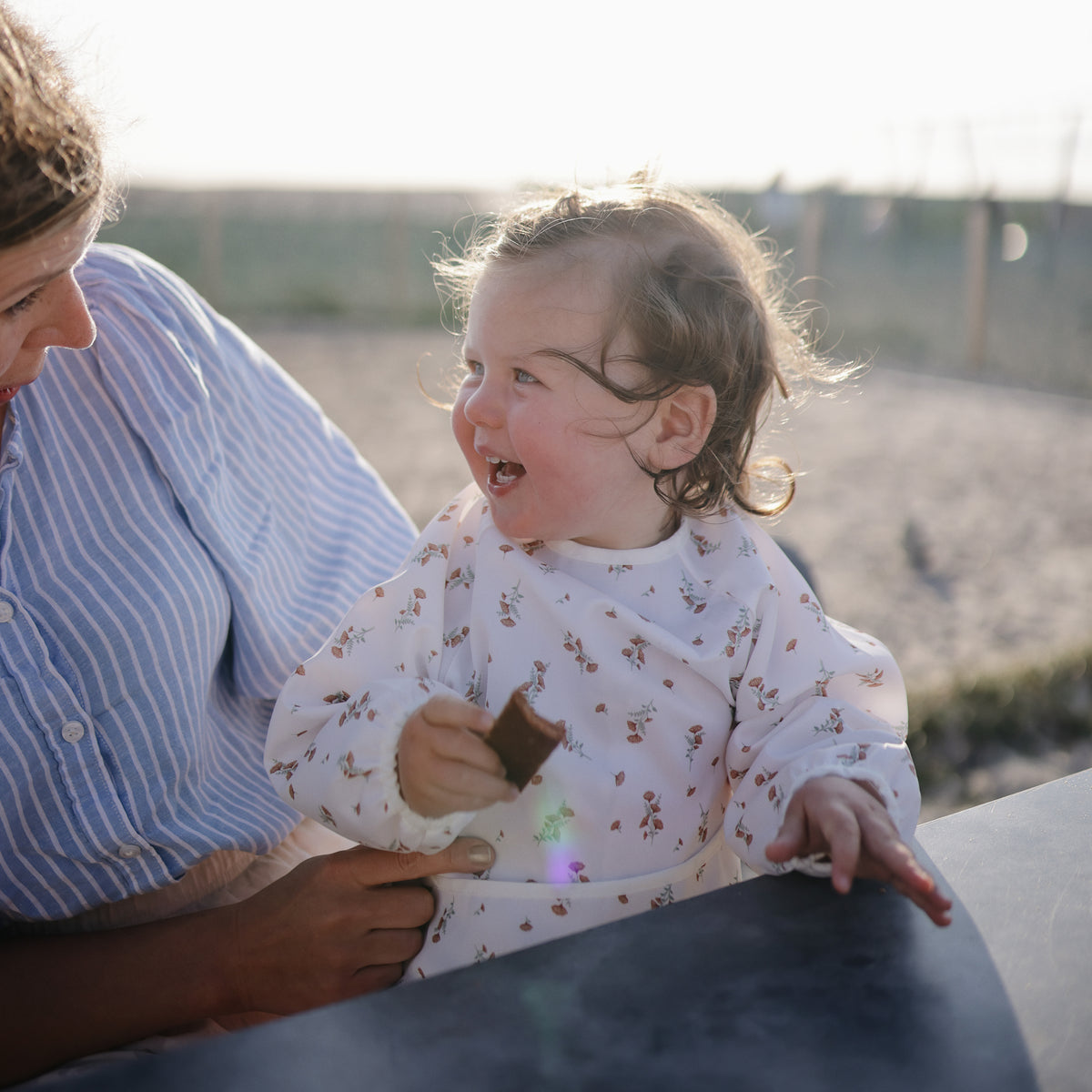 A toddler wearing a mushie Long Sleeve Bib holds food while sitting with an adult.
