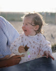 A toddler wearing a mushie Long Sleeve Bib holds food while sitting with an adult.