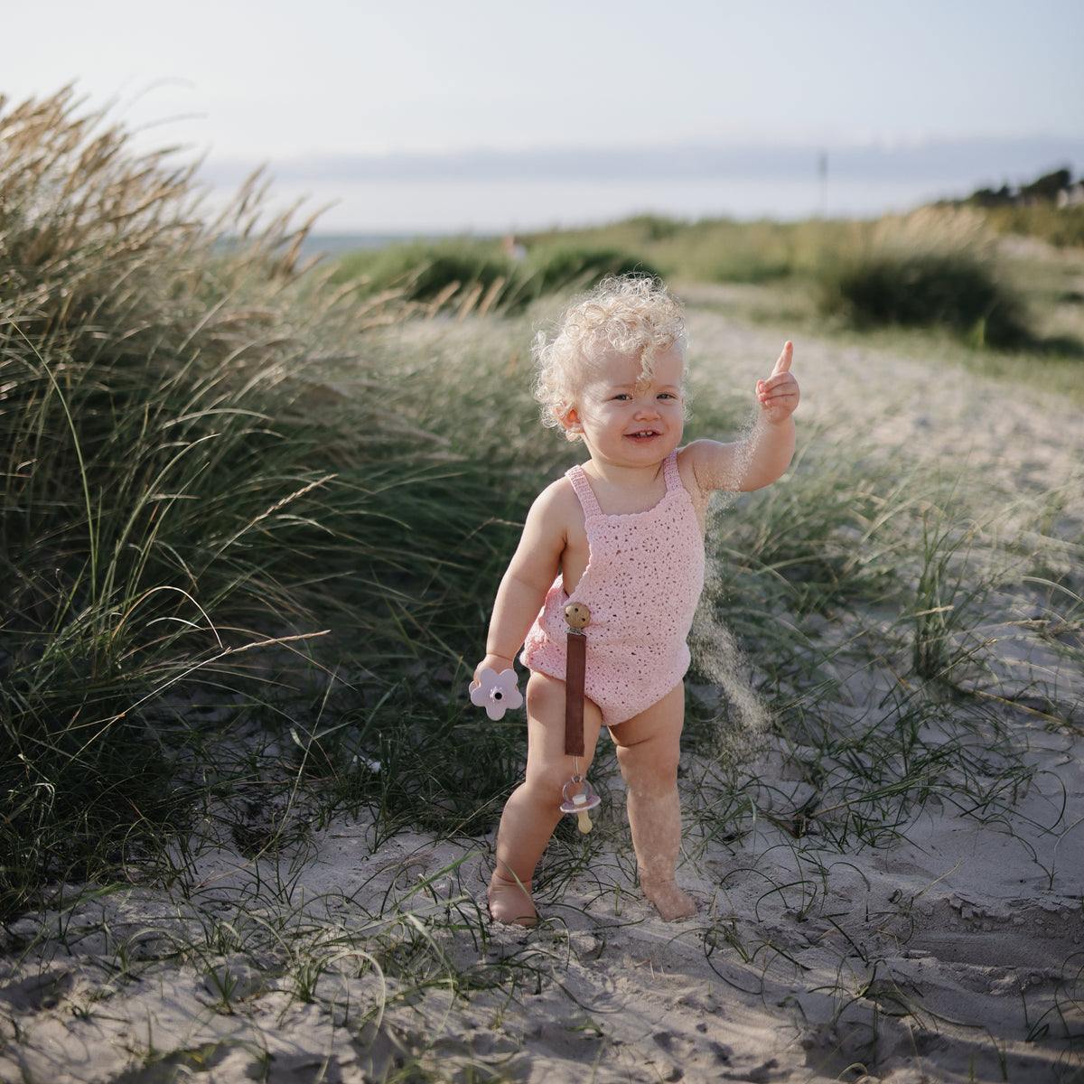 Smiling toddler with mushie Linen Pacifier Clip 2-Pack stands on sandy path, pointing up.
