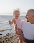 Smiling man holds baby with mushie Linen Pacifier Clip 2-Pack at the beach in sunlight.