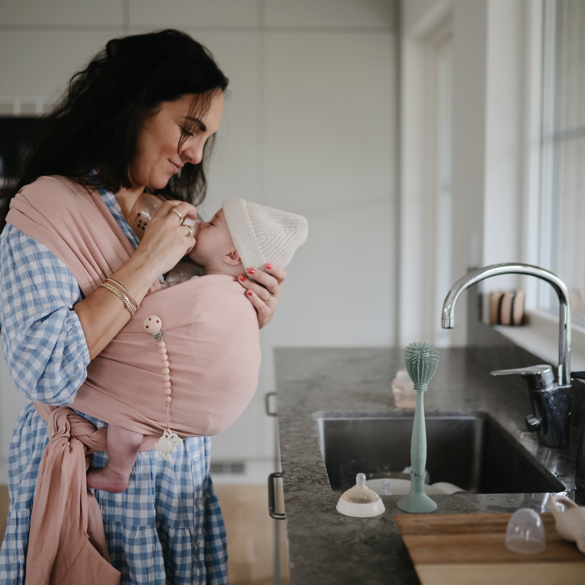 A smiling woman in a checkered dress cradles her baby in a pink sling in a modern kitchen. On the counter are FRIGG Butterfly Anatomical Silicone Pacifiers (0-6 Months, 2-Pack) and baby bottles beside the sink.