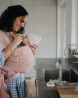 A smiling woman in a checkered dress cradles her baby in a pink sling in a modern kitchen. On the counter are FRIGG Butterfly Anatomical Silicone Pacifiers (0-6 Months, 2-Pack) and baby bottles beside the sink.