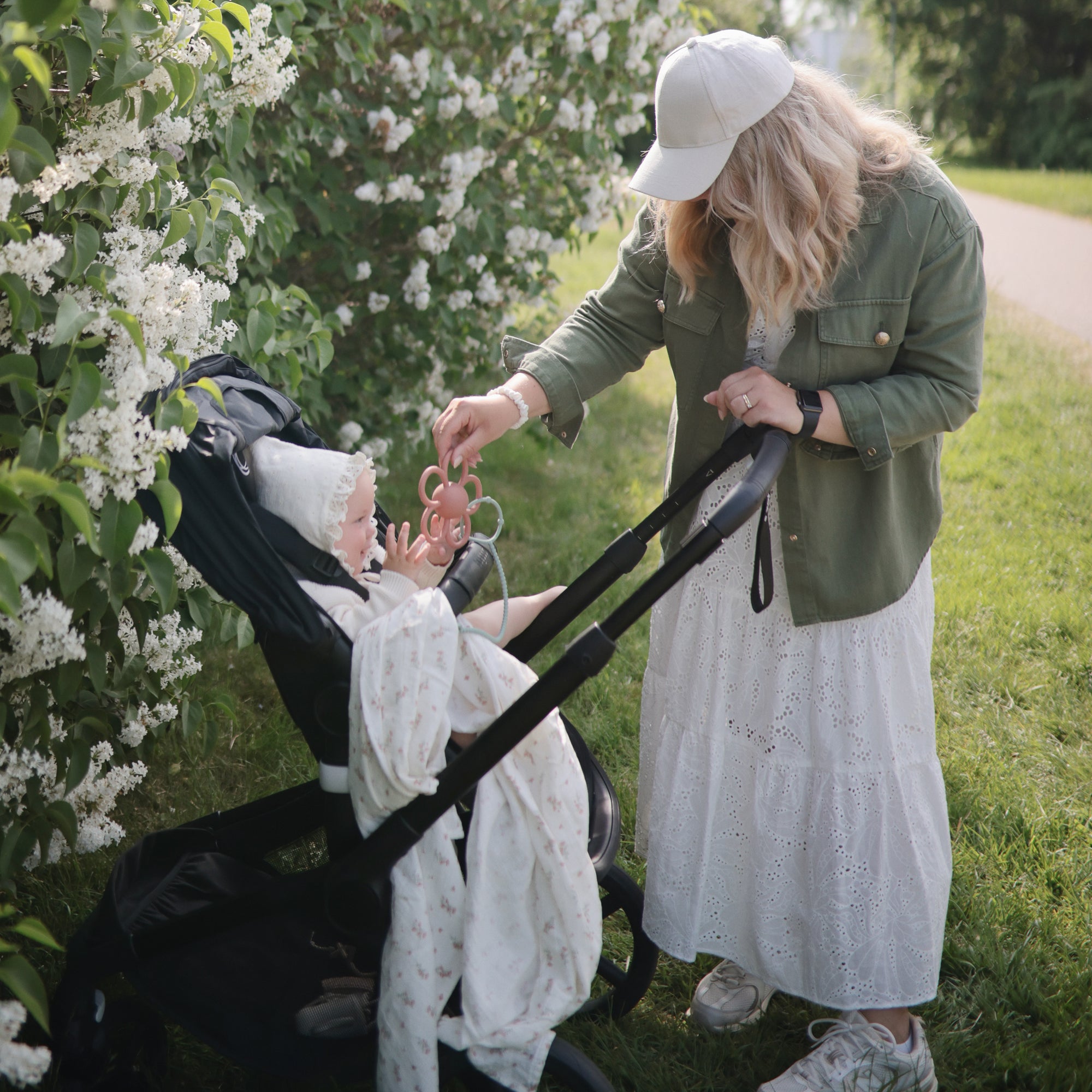 Woman gives mushie Organic Cotton Muslin Swaddle Blanket to baby in stroller by blooming bushes.