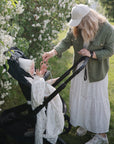 Woman gives mushie Organic Cotton Muslin Swaddle Blanket to baby in stroller by blooming bushes.