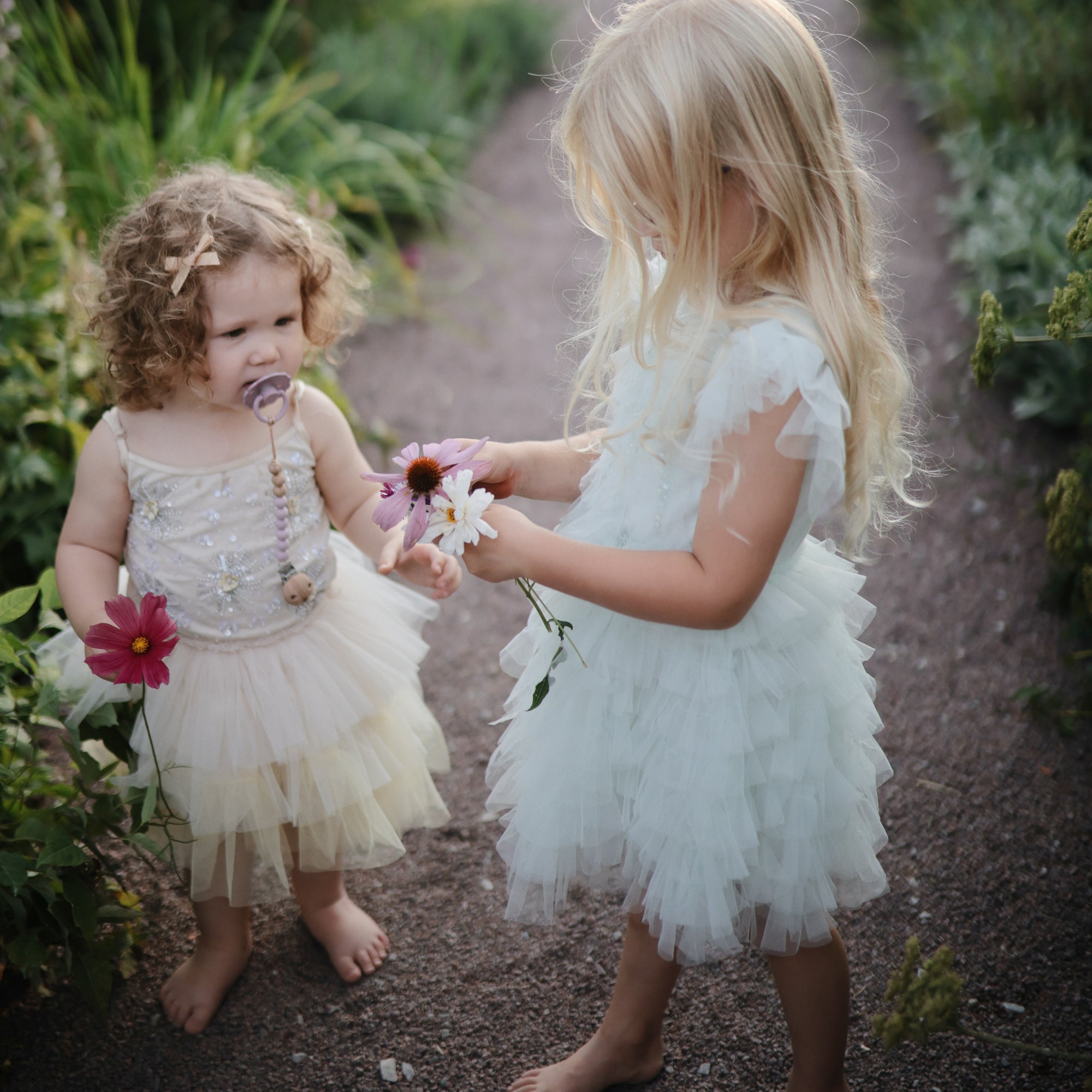 Two girls in frilly dresses stand barefoot, holding flowers and a FRIGG Daisy rubber pacifier.